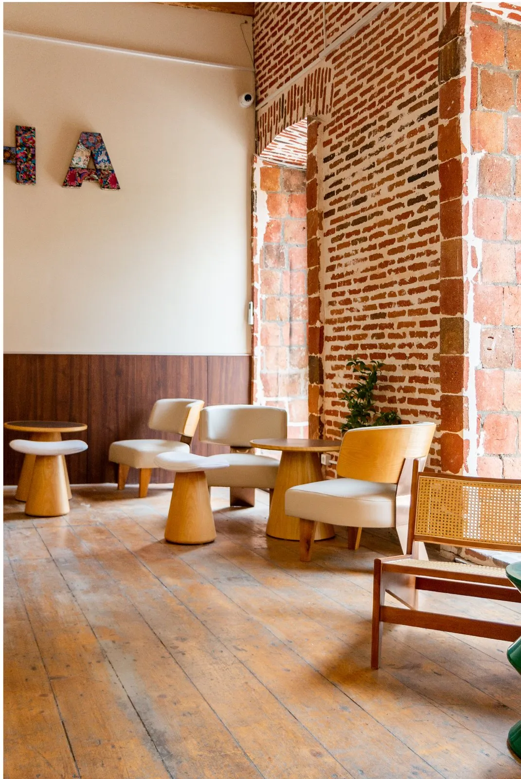 Timber armchairs and round tables beside an exposed brick wall and arched window